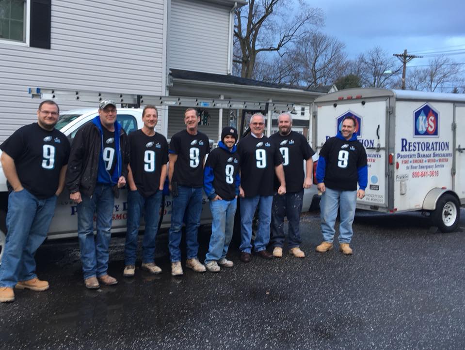 A group of eight people wearing matching number 9 jerseys stand in front of a K&S Restoration truck outside a building.