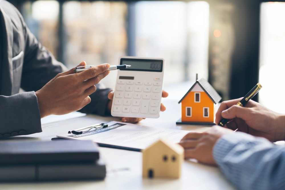 Two people discuss financial documents at a table with a calculator and model houses.